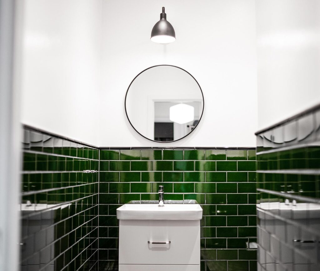 Renoverad modern toalett med mörkgrönt kakel, ett vitt handfat, en rund spegel och modern svart belysning