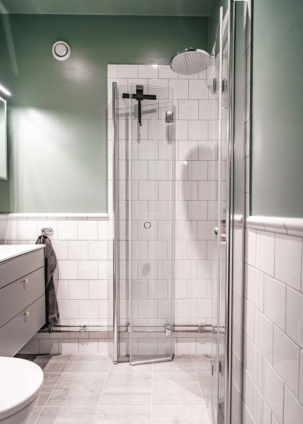 Modernt renoverat badrum med ljusgröna väggar, vitt kakel, ljusgrå klinkers, en dusch med ett vattenfallsmunstycke och modern belysning