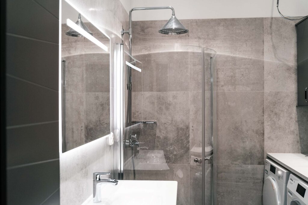 Nyrenoverat badrum med ljusgrå granit kakel på väggarna, en dusch med trekantigt munstycke, en stor spegel med belysning samt tvättmaskin och torktumlare