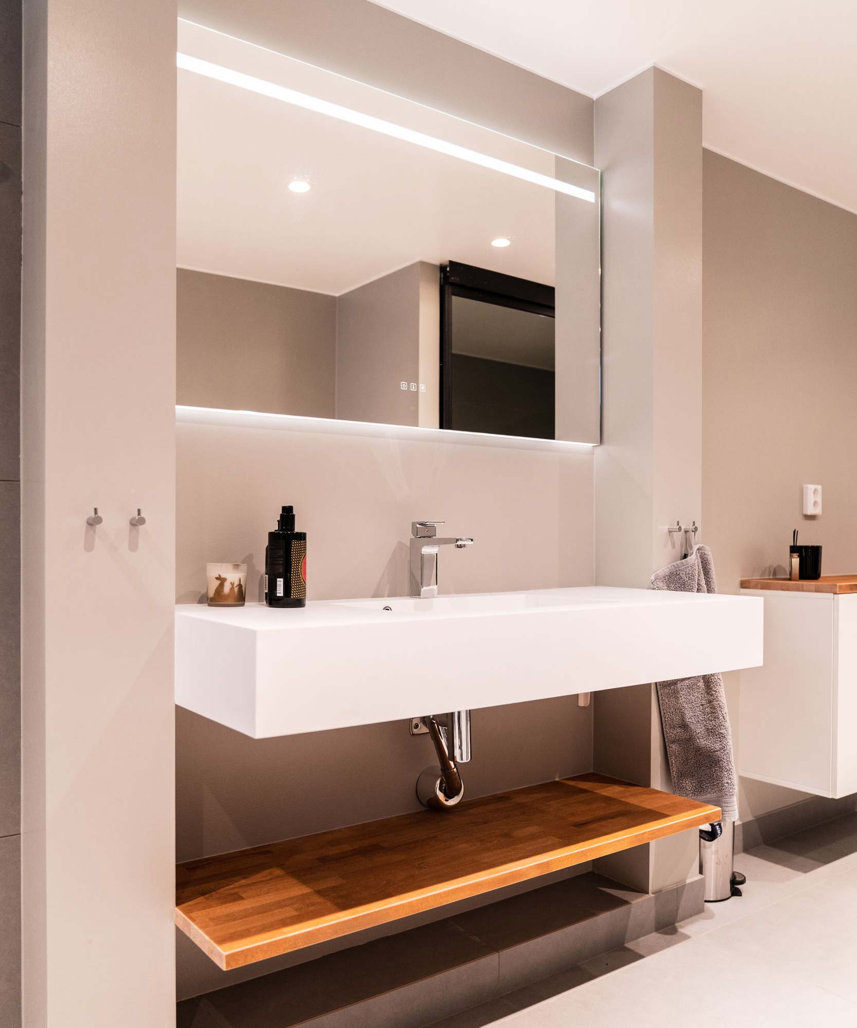 Lyxigt badrum med beiga väggar, ett stort vitt avlångt handfat med kantiga hörn samt en stor spegel med inbyggd belysning
