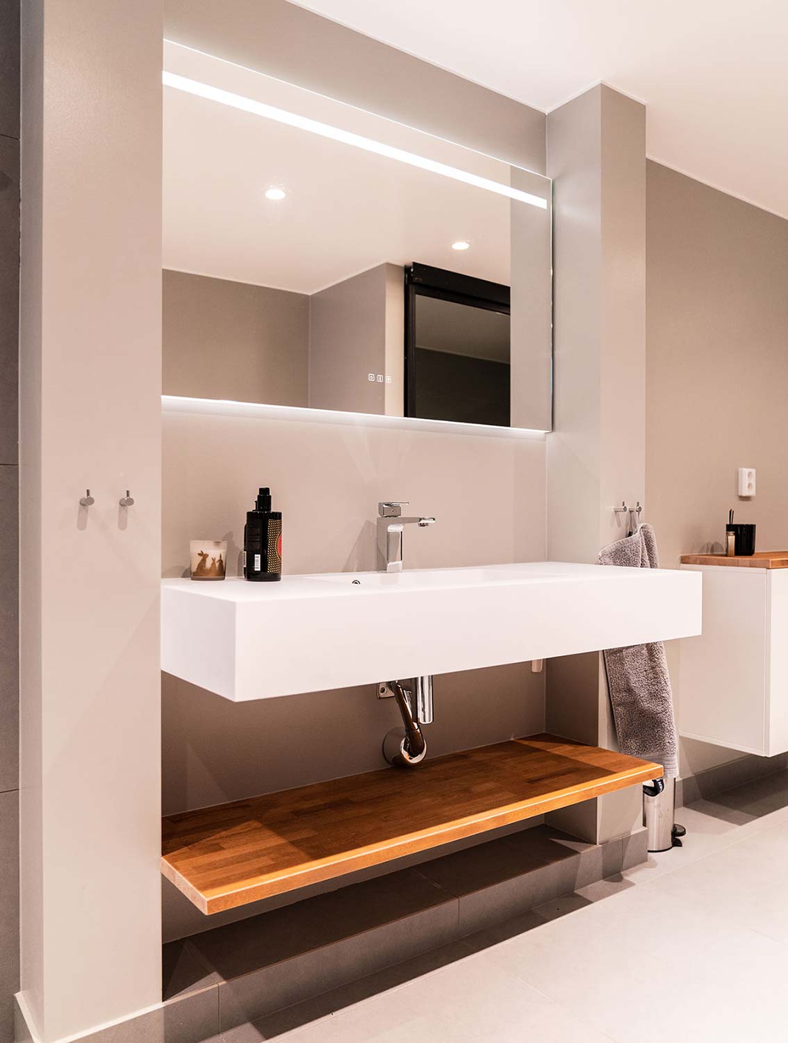 Lyxigt badrum med beiga väggar, ett stort vitt avlångt handfat, en stor spegel med inbyggd belysning och ett avlastningsbord under handfatet i trä