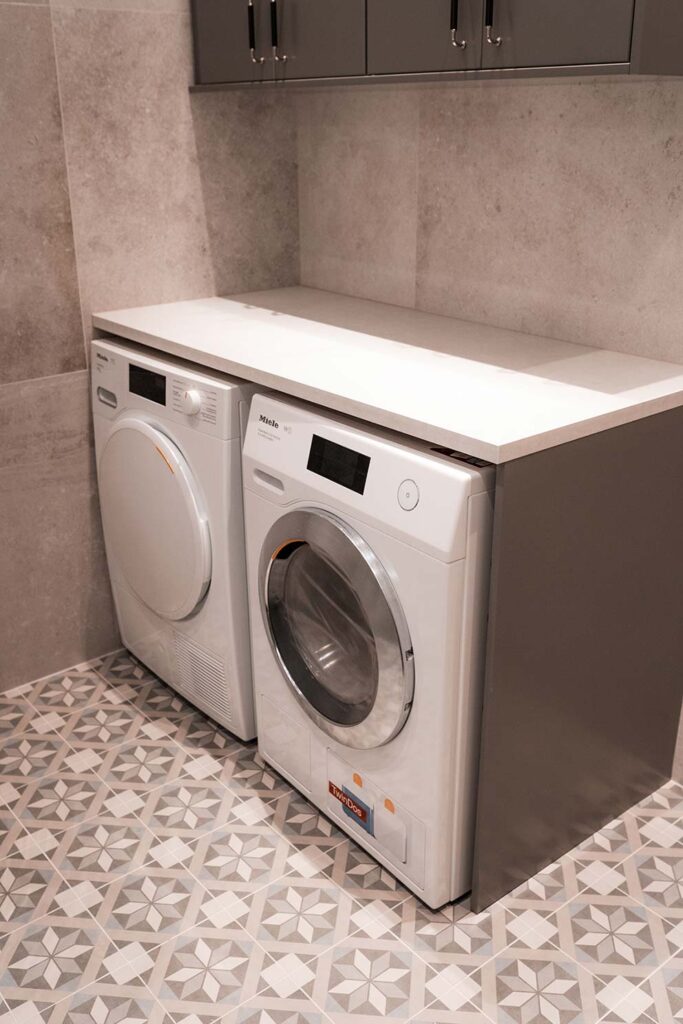 Tvättstuga med inbyggd tvättmaskin och torktumlare samt ett grått skåp ovanför, kakel av ljusgrå granit på väggarn och mönstrat klinker med stjärnor på golvet