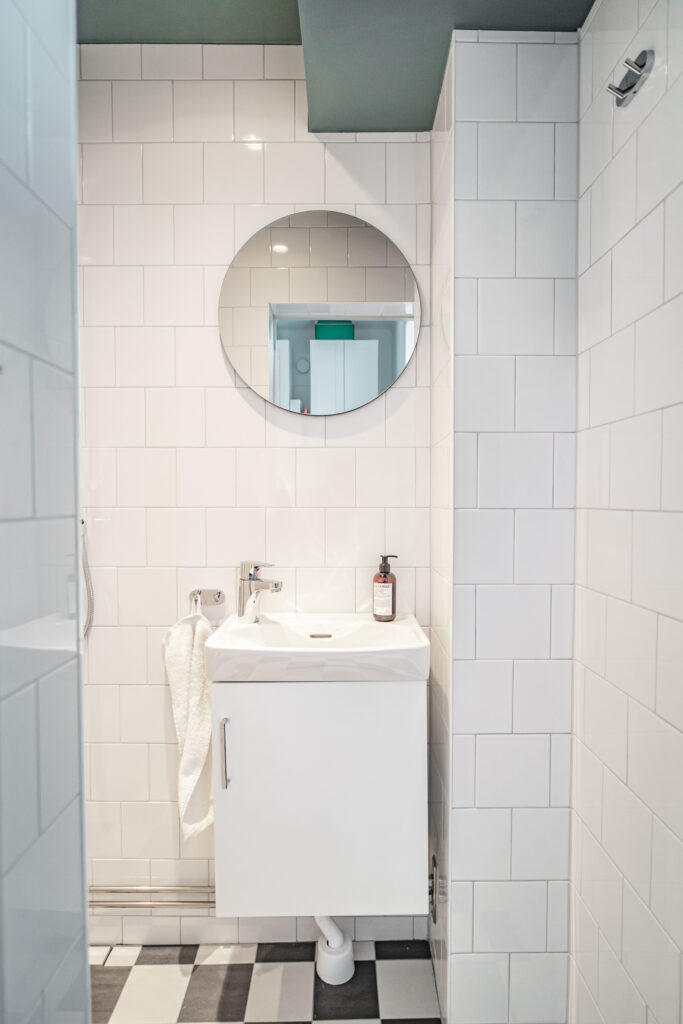 Toalett med vitt kakel på väggarna, ett vitt litet handfat och en rund spegel, modern belysning samt schackrutigt golv i svart och vitt