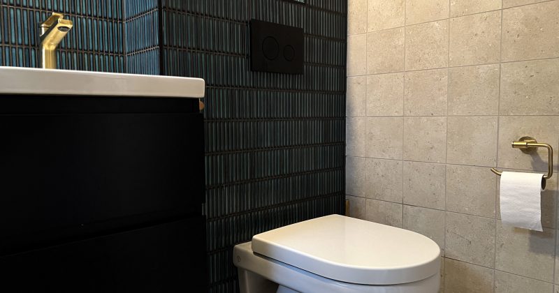 Toalett inbyggd i väggen, unikt kakel i turkost och guld på väggarna, en vattenkran i guld och ett svart badrumsskåp