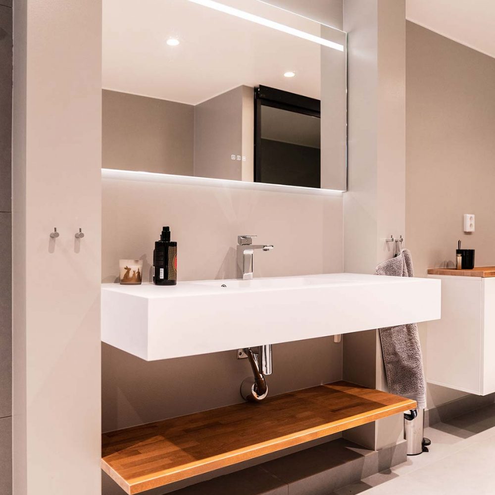 Lyxigt badrum med beiga väggar, ett stort vitt avlångt handfat, en stor spegel med inbyggd belysning och ett avlastningsbord under handfatet i trä