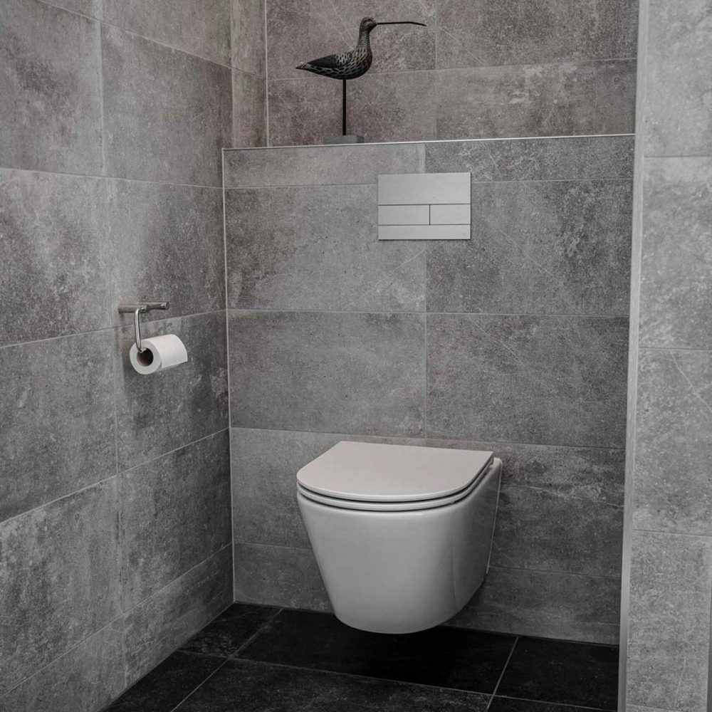 Badrum med kakel av ljusgrå granit på väggarna, svart klinker på golvet, en vit inbyggd toalett och en modern spolfunktion på väggen